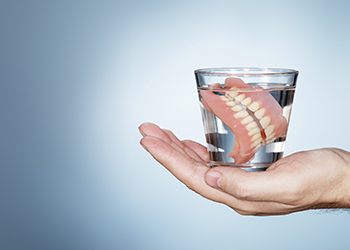 An image of a person holding a glass of water that contains a full set of dentures in the palm of their hand
