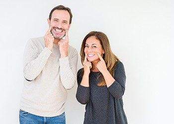 A middle-aged couple pointing to their smiles after undergoing in-practice teeth whitening